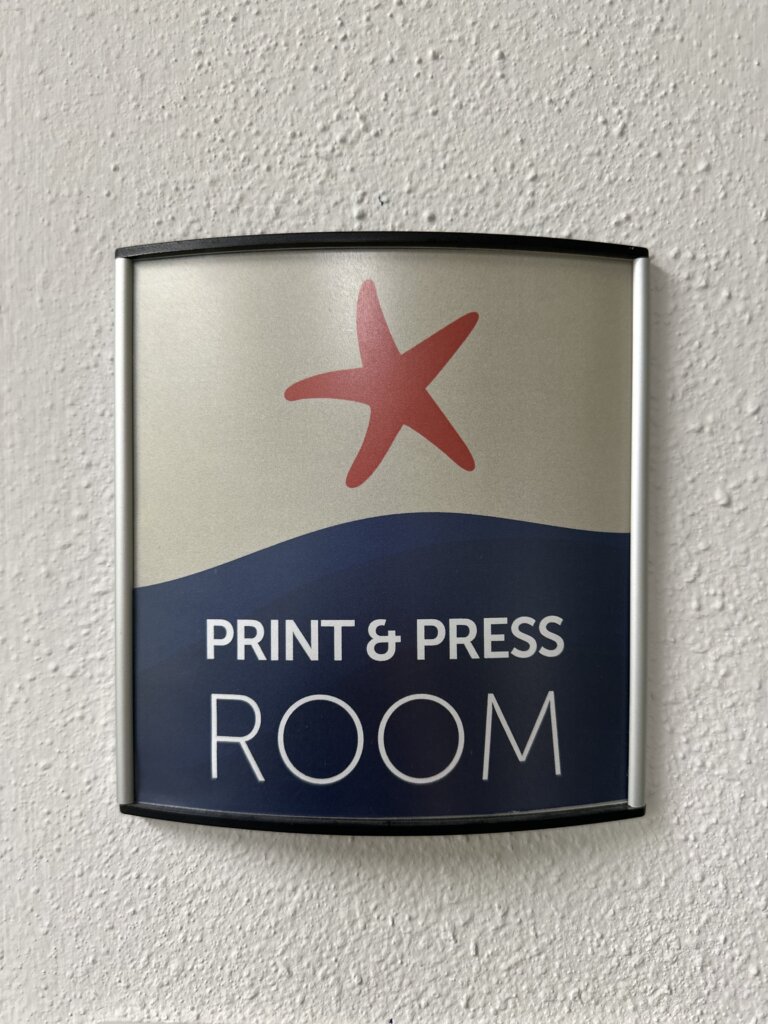 a photograph of a small wall sign - the top part of the sign has a red starfish, and the lower half is navy blue. White words read "Print & Press Room"