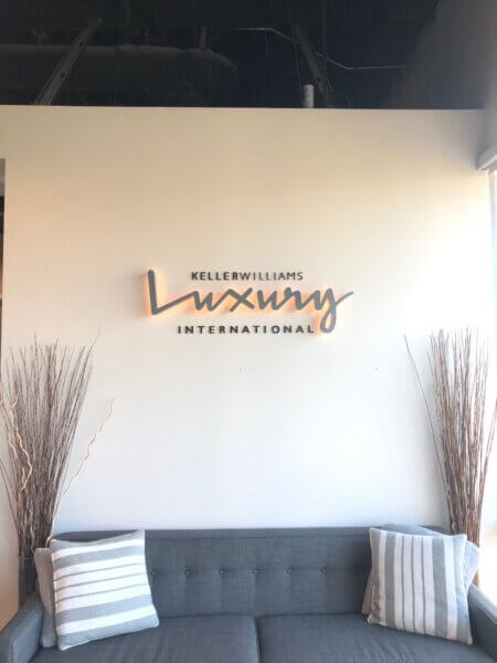 Picture shows a white wall (with a gray couch set against it) - on the wall is a mounted illuminated sign that reads "KellerWilliams Luury International"