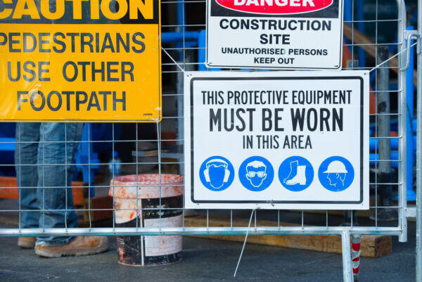 Warning signs on fence at construction site with symbols for compulsory protective gear to enter, legs of worker in boots in background.