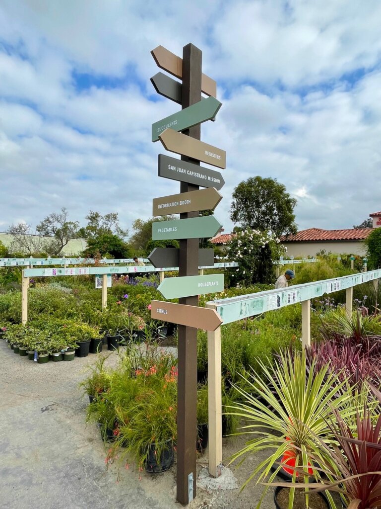 A photo of a navigation sign in Plant Depot's outside yard; arrows point in different directions to guide guests to different buildings or shopping sections (ex. "Vegetables")